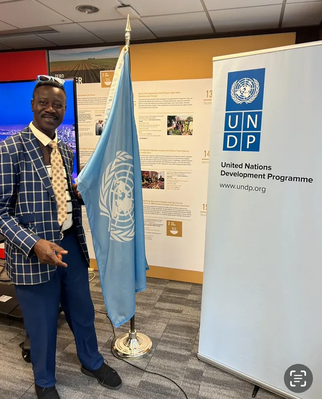 Chuck Eke standing beside a United Nations flag at a United Nations Development Programme stand